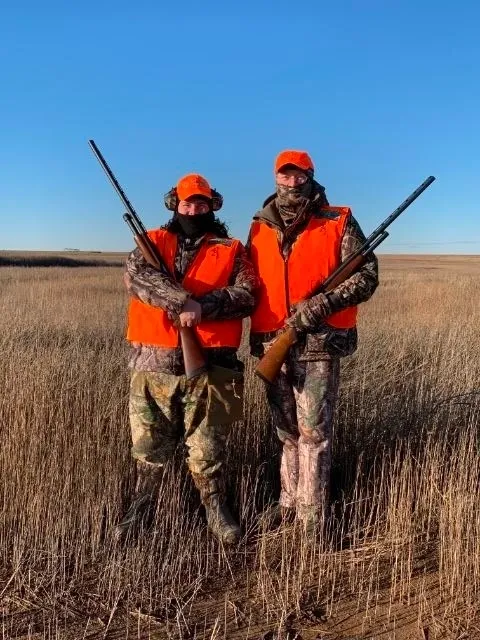 Two hunters in orange vests holding guns standing on a dry grass field.