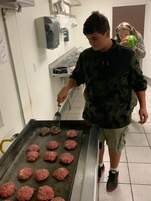 A boy is cooking hamburgers on an outdoor grill.