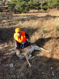 A man in an orange vest is sitting on the ground with a dead animal.