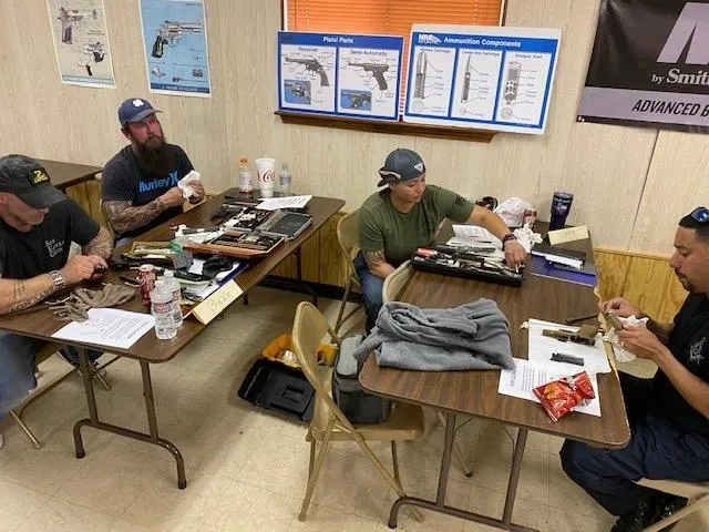 A group of men sitting at tables in front of some guns.