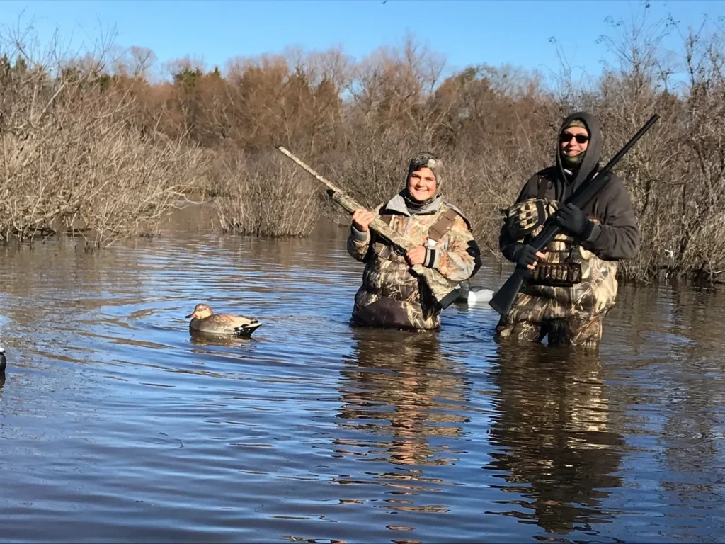 Two people in the water with guns and a dog.