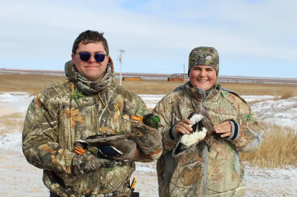 Two people in camouflage holding ducks and wearing sunglasses.