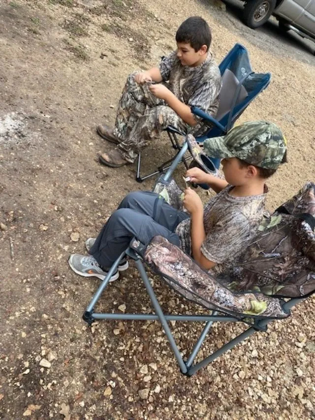 Two young boys sitting in camo chairs on a gravel ground.