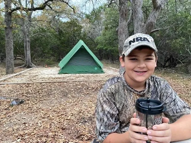 A boy in camouflage holding a cup near a tent.