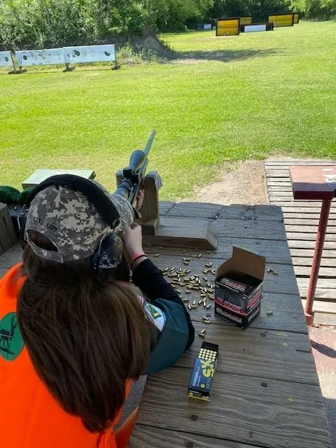 A woman is sitting on the ground shooting a rifle.