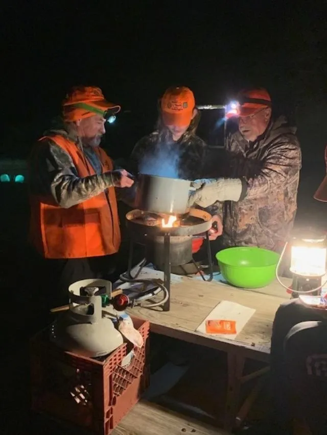 Three men in orange jackets cooking food on a fire.