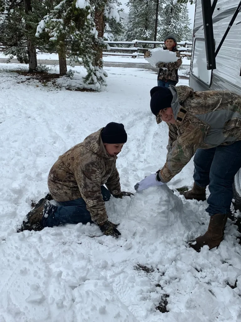 Two men in camouflage jackets and hats building a snowman.
