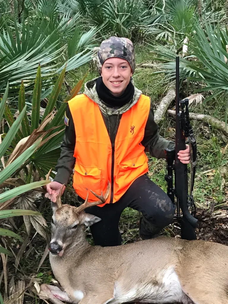 A woman in an orange vest holding a rifle and standing next to a deer.