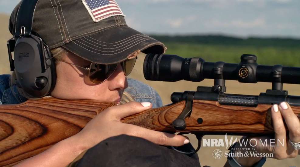 A woman in sunglasses and hat looking through the scope of her rifle.